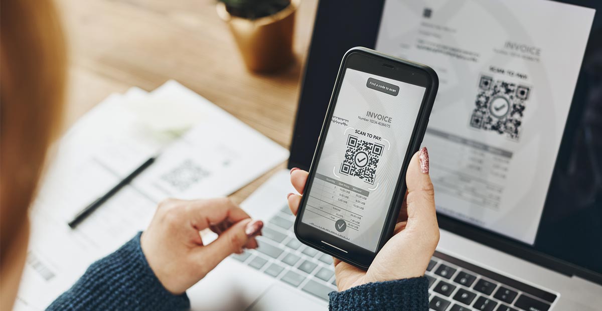 Die E-Rechnung Eine Frau halt ein Smartphone vor einen Laptop und fotografiert einen QR-Code ab